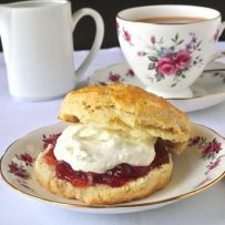 A flowered plate with a scone, jam and clotted cream and a cup of tea in the background