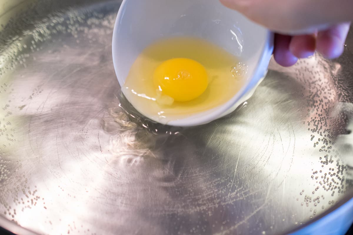 Pouring an egg from a white bowl into a pan of simmering water 