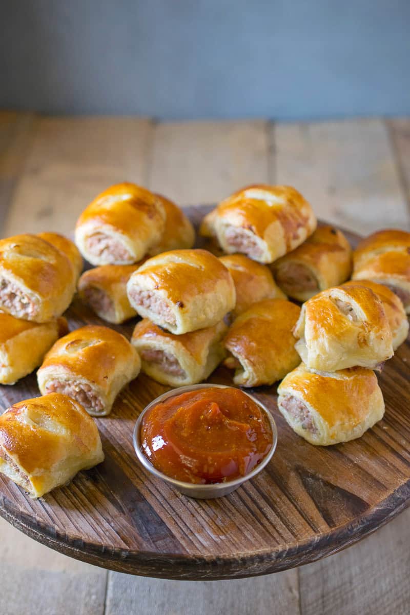 Mini pork  apple & sage sausage rolls are stacked on a wooden serving board with a silver bowl of curry ketchup