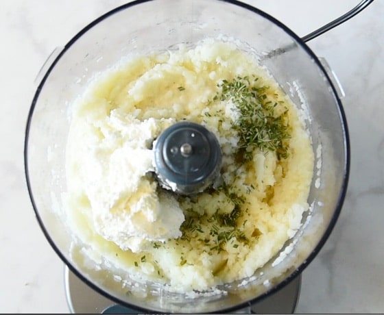 Adding cream cheese, rosemary, salt and pepper to a food processor of blended cauliflower