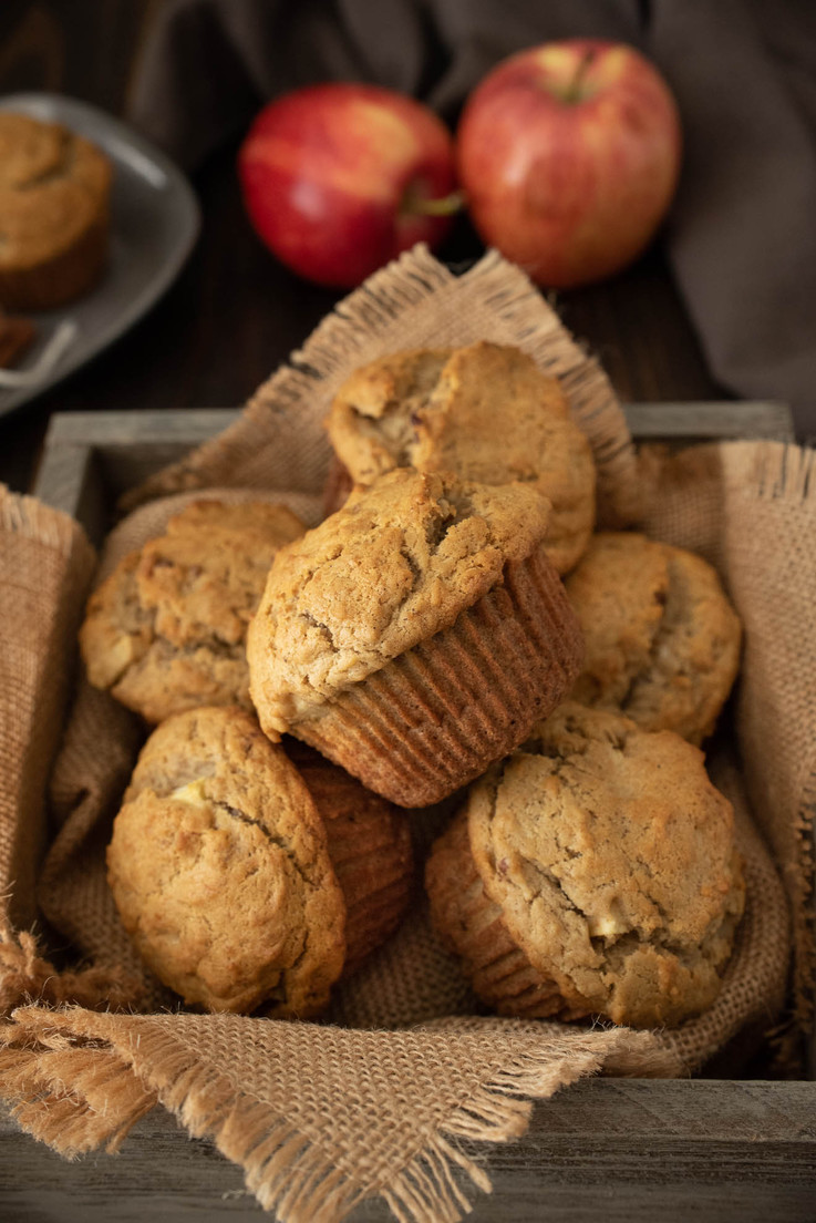 A wooden box full of muffins with red apples in the background