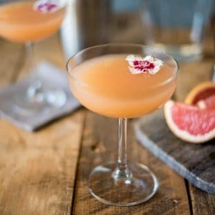 A coupe champagne glass of April Showers cocktail with a flower and grapefruit wedges