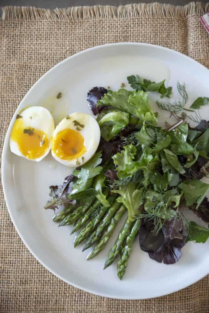A closeup of a green salad mixed with fresh herbs, fresh asparagus and a boiled egg cut in half.