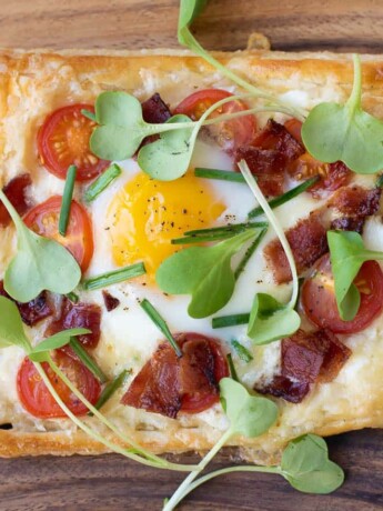 A square of puff pastry topped with cheese, tomatoes, bacon, egg, chives and greens