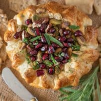 Vibrant pomegranate seeds with rosemary leaves top a baked brie that is wrapped in puff pastry served with a knife