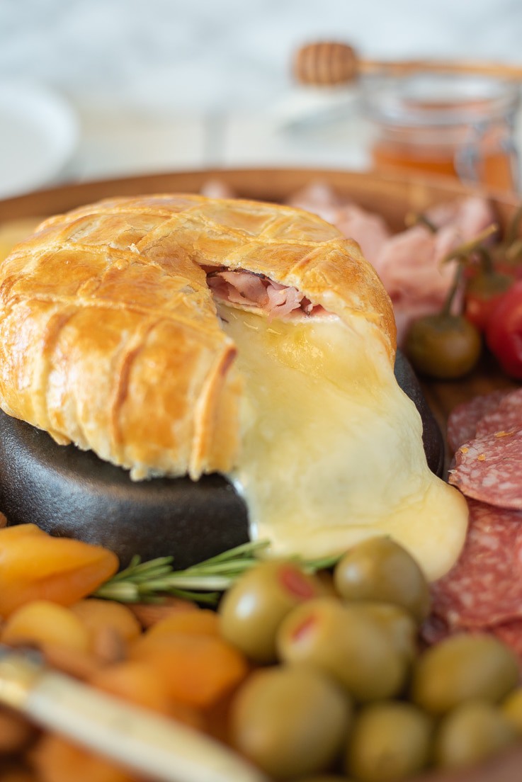 A wheel of Baked Brie and Black Forest ham in puff pastry is fresh out of the oven and the warm melted brie is running all over a charcuterie board