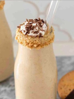 Tall glass of banoffee pie milkshake topped with whipped cream, sliced fresh bananas, crushed graham crackers, and a rich drizzle of dulce de leche.