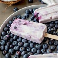 A closeup of a blueberry coconut frozen yogurt popsicle on top of fresh blueberries