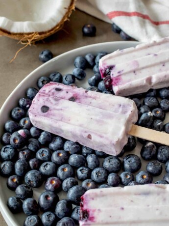A closeup of a blueberry coconut frozen yogurt popsicle on top of fresh blueberries