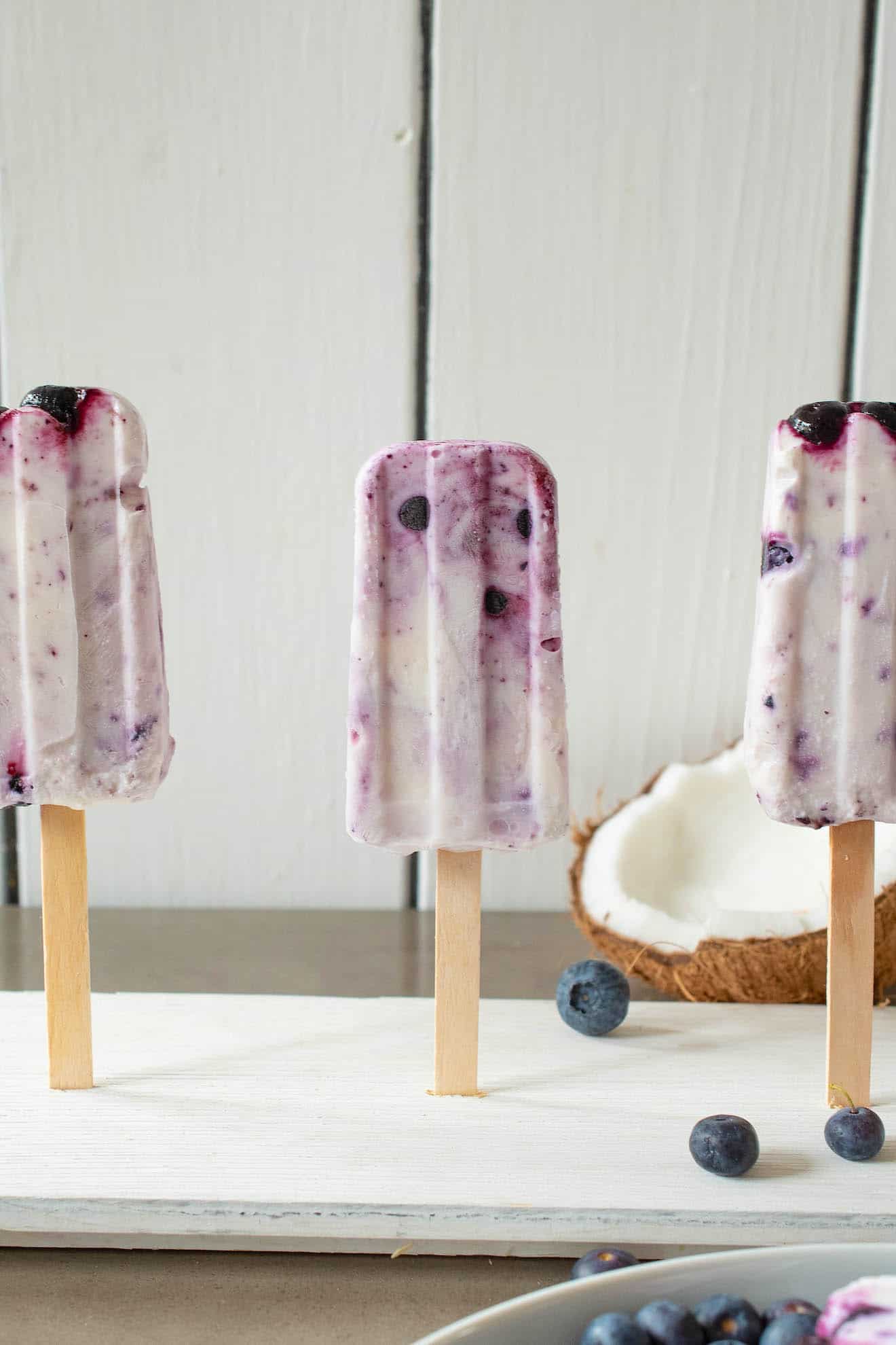3 blueberry coconut frozen yogurt popsicles standing up with a real coconut and blueberries