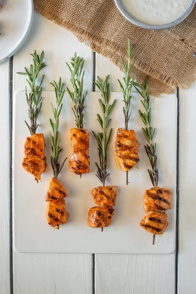 Rosemary skewered chicken served on a white platter with dipping sauce viewed from overhead