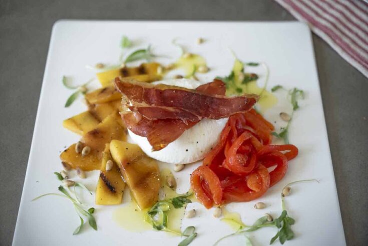 Creamy burrata surrounded by sliced peach, red pepper strips and prosciutto