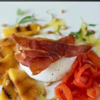 A ball of burrata topped with prosciutto that has been crisped in the oven, served with red pepper strips and sliced peaches