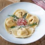 A round white pasta bowl with 4 butternut squash filled cappellacc with a small amount of marinara sauce in the center with crispy sage leaves and grated Parmesan cheesei
