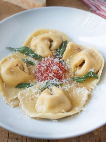 A round white pasta bowl with 4 butternut squash filled cappellacc with a small amount of marinara sauce in the center with crispy sage leaves and grated Parmesan cheesei