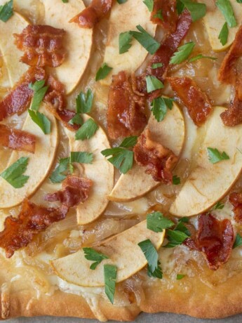 A closeup of the topping on a flatbread pizza of fontina cheese, apple, caramelized onions, bacon pieces and green parsley