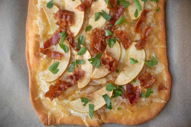A closeup of the topping on a flatbread pizza of fontina cheese, apple, caramelized onions, bacon pieces and green parsley