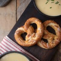 2 bowls of cheese and beer soup served with 2 soft pretzels on a board with a red and white striped napkin