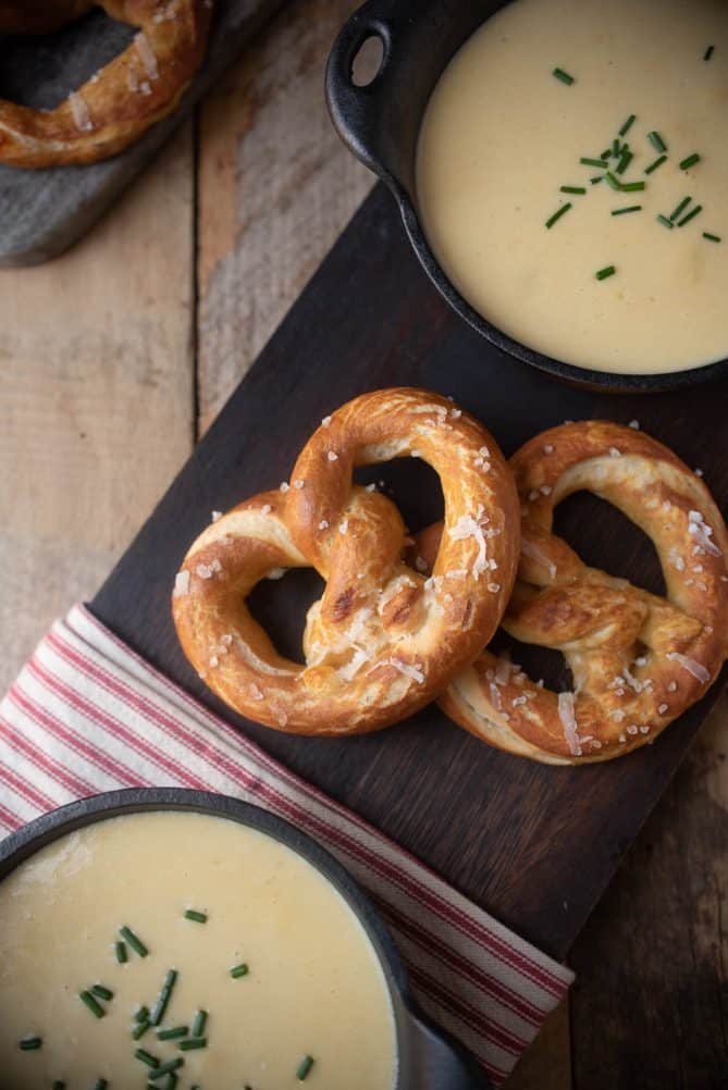 2 bowls of cheese and beer soup served with 2 soft pretzels on a board with a red and white striped napkin