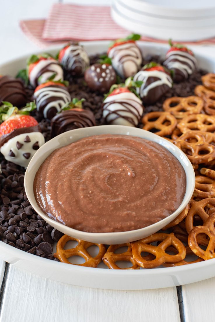 Chocolate covered strawberry dessert hummus in a white bowl with chocolate covered strawberries and pretzels