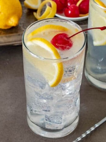 A tall glass filled with ice and Tom Collins garnished with lemon slices and a cherry