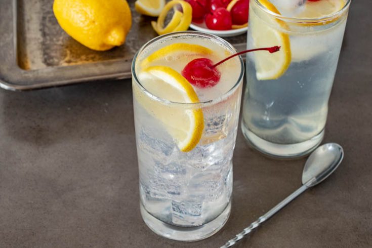 A tall glass filled with ice and Tom Collins garnished with lemon slices and a cherry