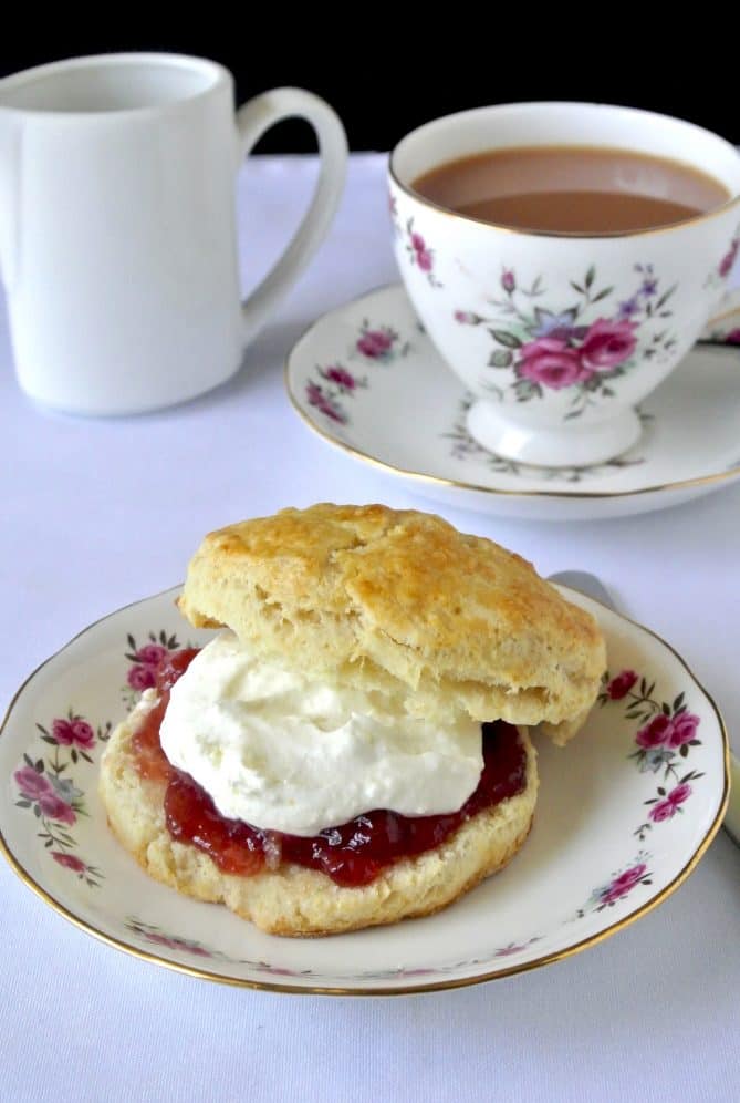 A scone cut in half topped with jam and clotted cream with a flower decorated china tea cup filled with tea