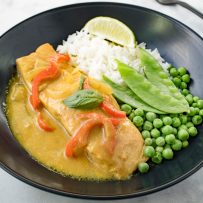 A filet of salmon bathed in coconut curry sauce with fresh peas, snow peas and white rice in a black bowl