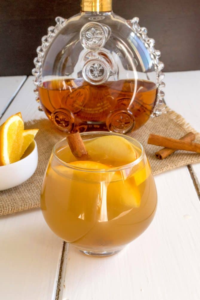 A short glass of Cognac apple cider garnished with orange wedges, apple wedges and a cinnamon stick
