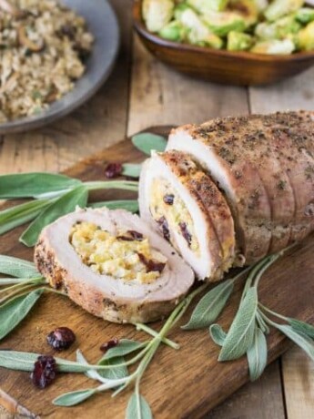 Apple cranberry cornbread stuffed pork loin served on a cutting board garnished with fresh sage leaves with a bowl of Brussels sprouts and rice in background