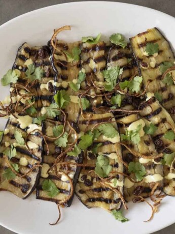 Slices of aubergine/eggplant with grill marks on a plate topped with curry yogurt sauce, crispy onions, raisins and coriander/cilantro.