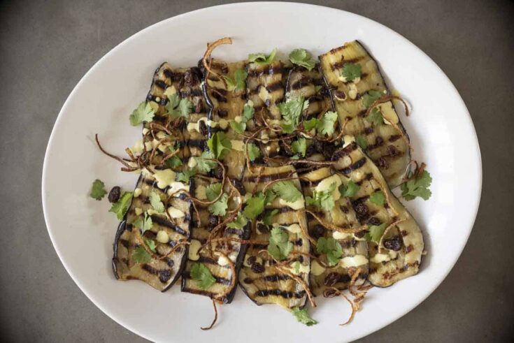 Slices of aubergine/eggplant with grill marks on a plate topped with curry yogurt sauce, crispy onions, raisins and coriander/cilantro.