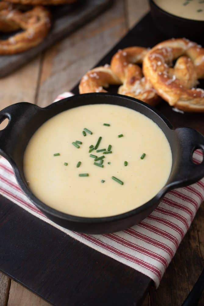 Creamy off white beer cheese soup in a black bowl topped with chives with pretzels in the background