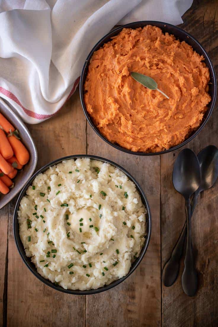 Mashed russet potatoes with melted butter and chives and mashed sweet potatoes both in black bowls with serving spoons