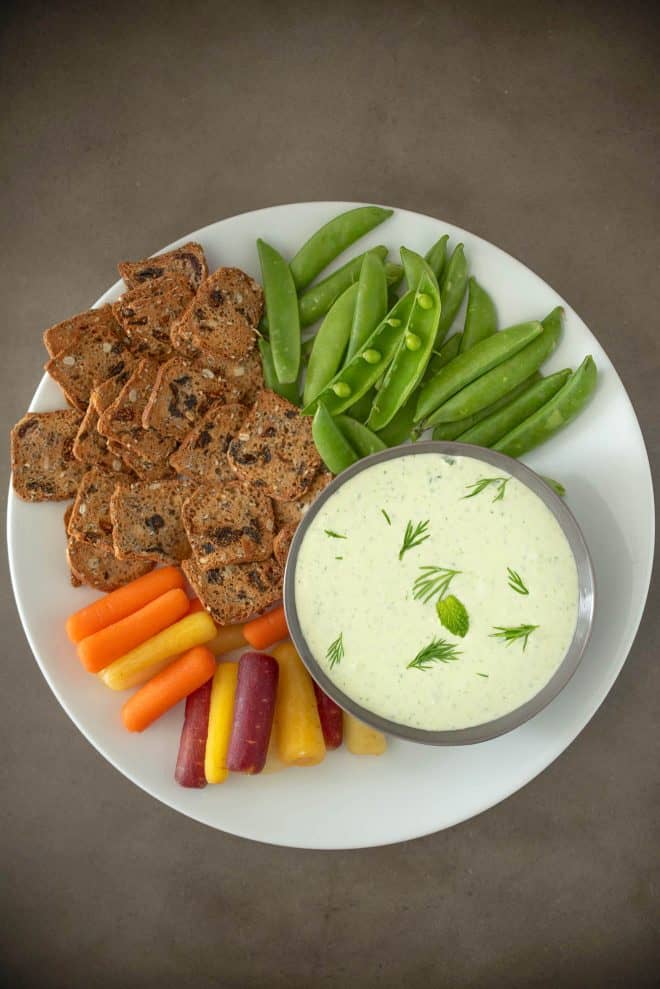 Tzatziki dipping sauce in a grey bowl on a white plate surrounded by vegetables and crackers.