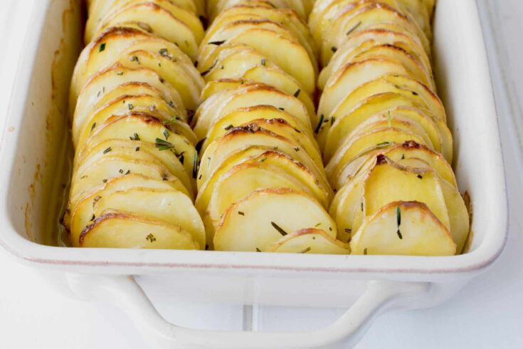 A closeup of thinly sliced potatoes stacked together vertically topped with rosemary