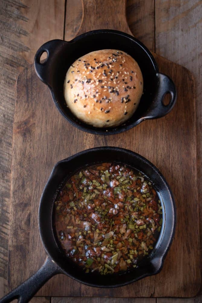 A small cast iron bowl that holds a crusty bread roll alongside a cast iron skillet with bacon, sage butter