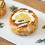 A white serving plate with crab cakes topped with lemon scallion sauce, lemon and fresh dill