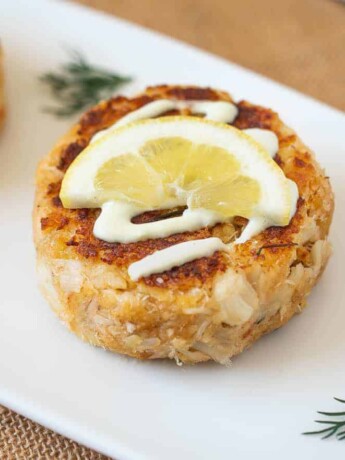 A white serving plate with crab cakes topped with lemon scallion sauce, lemon and fresh dill