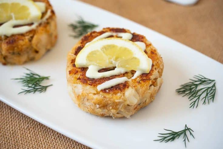 A white serving plate with crab cakes topped with lemon scallion sauce, lemon and fresh dill