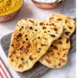 Two pieces of golden, blistered yeast-free homemade naan bread served with a bowl of lentil dal and rice in the background.