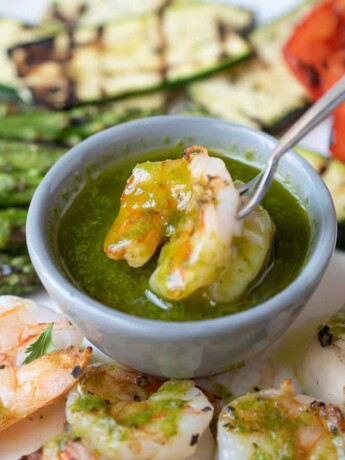 Herb sauce in a grey bowl with a shrimp on a fork dipping it into the sauce surrounded by more shrimp and grilled vegetables