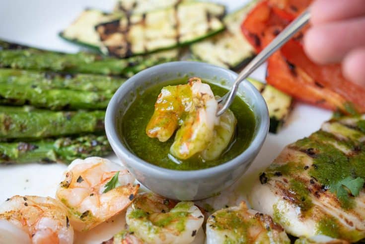 Green herb sauce in a small bowl with a shrimp on a fork being dipped into it surrounded by vegetables