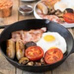 Fried egg, bacon, sausage, tomato, mushrooms in a cast iron pan.
