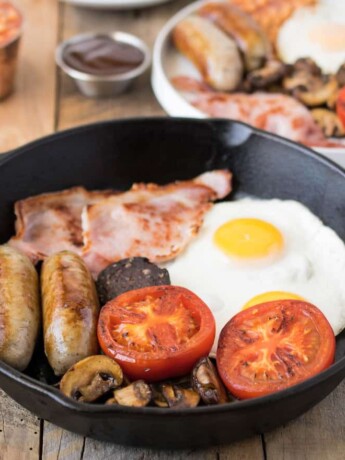 Fried egg, bacon, sausage, tomato, mushrooms in a cast iron pan.