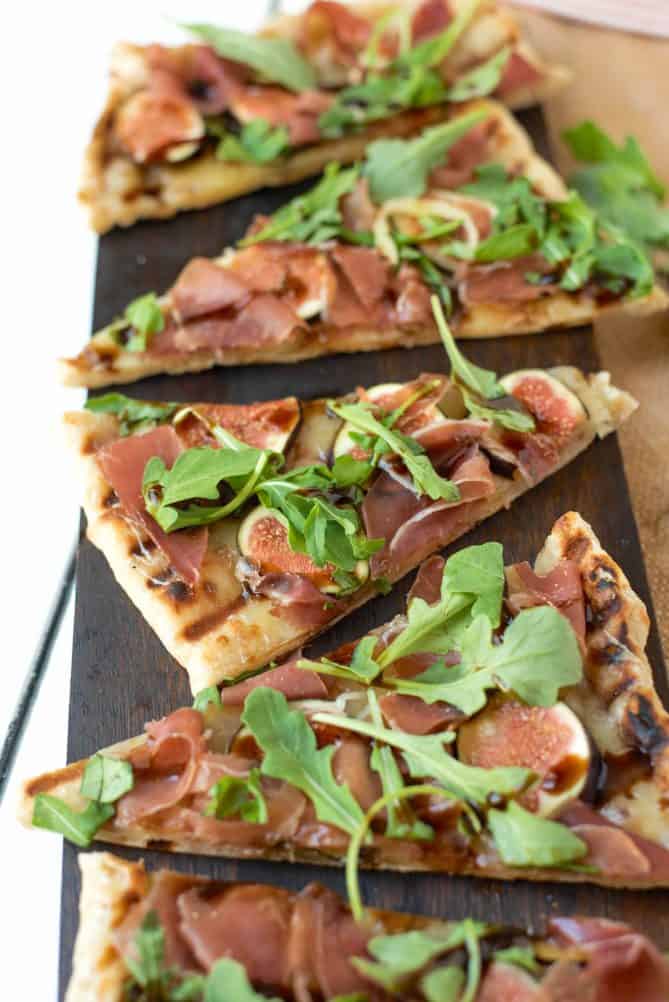 Triangle slices of grilled flatbread with figs, prosciutto and arugula on a wood serving board