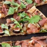 A slice of grilled flatbread topped with fontina, sliced fresh figs, prosciutto, arugula leaves and balsamic glaze