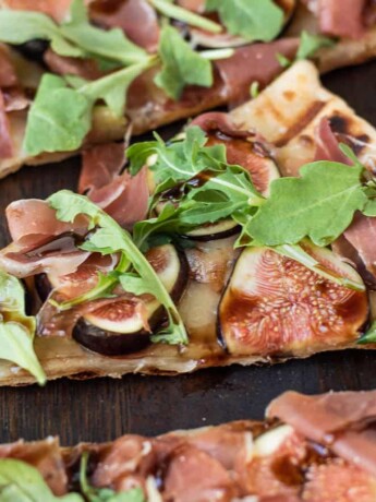 A slice of grilled flatbread topped with fontina, sliced fresh figs, prosciutto, arugula leaves and balsamic glaze