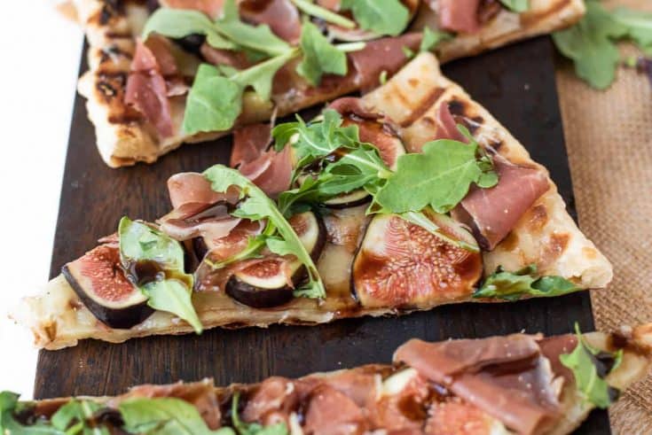 A slice of grilled flatbread topped with fontina, sliced fresh figs, prosciutto, arugula leaves and balsamic glaze