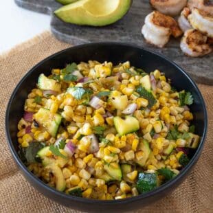 A black bowl filled with corn kernels and zucchini with a serving spoon and grilled shrimp